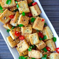 Close-up top view of a plate with fried tofu cubes garnished with chopped spring onion, red chili, and toasted white sesame seeds.