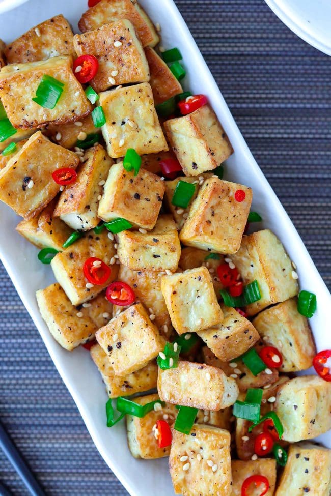 Close-up top view of a plate with pan-fried tofu cubes garnished with chopped spring onion, red chili, and toasted white sesame seeds.