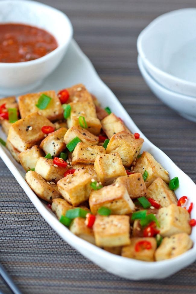 Front view of a long white plate with a bowl of spicy dipping sauce and crispy pan-fried tofu cubes sprinkled with chopped spring onion, chopped red chili, and toasted white sesame seeds. Small plates on right side of the plate with tofu.