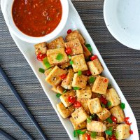 A long white plate with a bowl of spicy dipping sauce and pan-fried tofu cubes sprinkled with chopped spring onion, chopped red chili, and toasted white sesame seeds. Black chopsticks and small plates on either side of the plate.