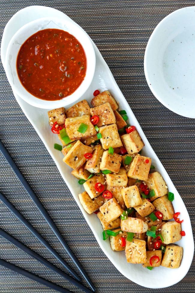A long white plate with a bowl of spicy dipping sauce and pan-fried tofu cubes sprinkled with chopped spring onion, chopped red chili, and toasted white sesame seeds. Black chopsticks and small plates on either side of the plate.