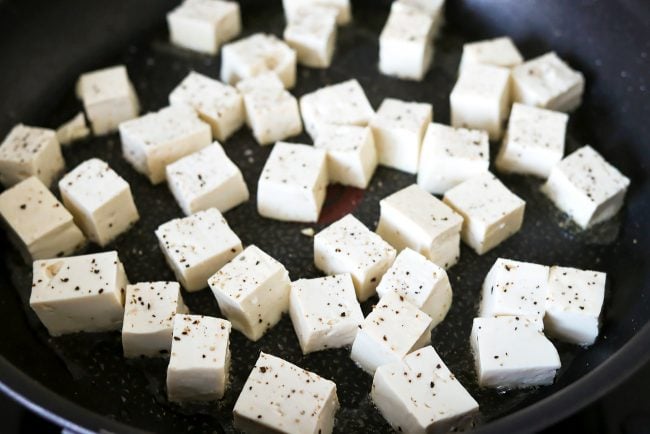 Tofu cubes seasoned with salt and pepper in a black nonstick skillet with a bit of oil.