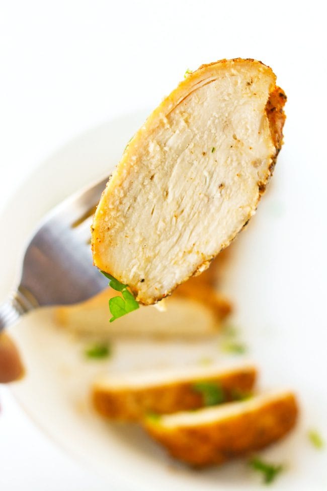 Fork holding up a piece of chicken above a plate with a sliced baked chicken breast that is garnished with chopped fresh parsley.
