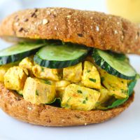 A Mango Chicken Salad Bagel Sandwich with mixed salad greens and cucumber slices on a white plate.
