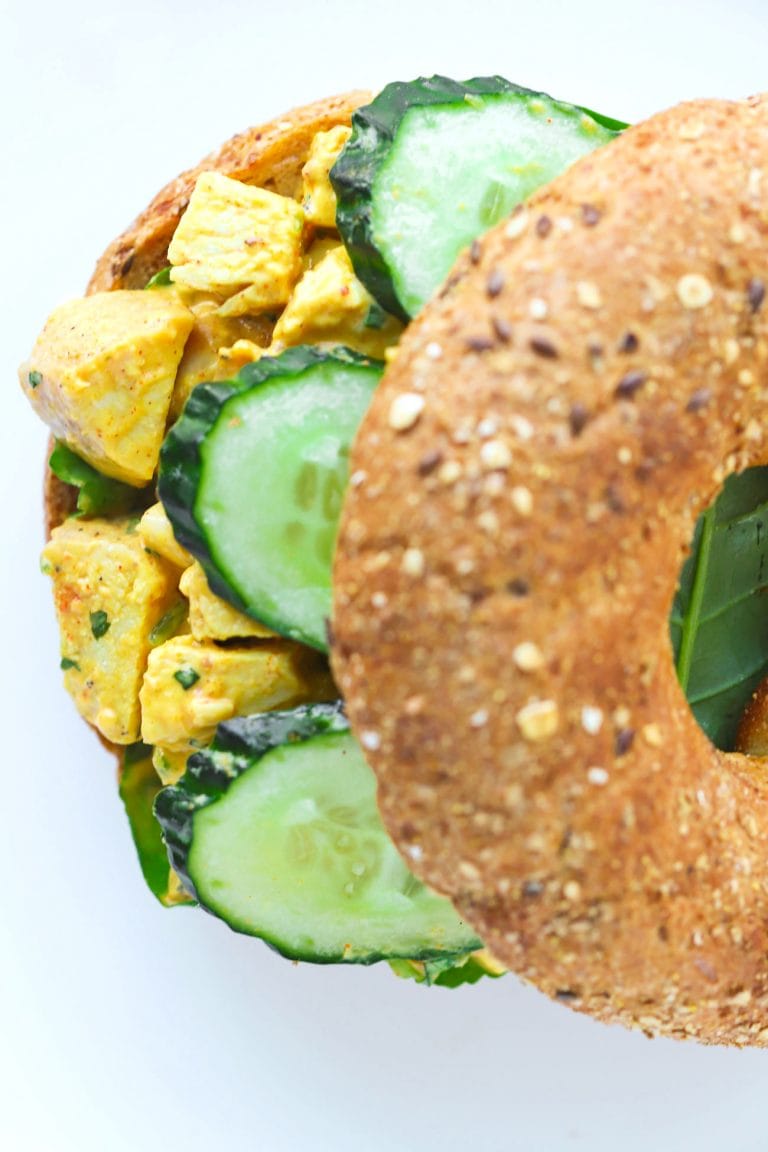 Top view of a mango chicken salad bagel sandwich with spinach leaves and cucumber slices on a white plate.