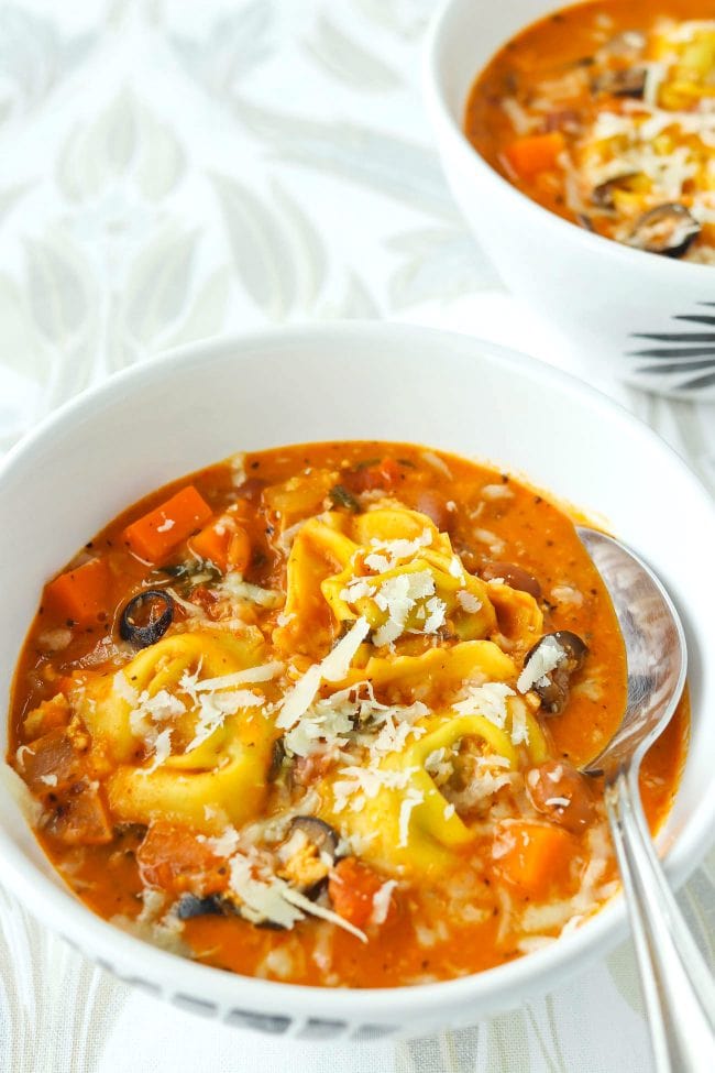 Front view of two diagonally placed bowls of creamy tomato basil soup with tortellini, ground chicken, pinto beans, diced carrot, and sliced black olives topped with grated cheese. Front bowl has a silver spoon.