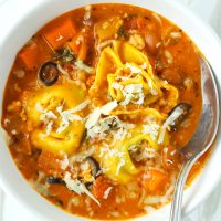 Tomato basil tortellini soup with pinto beans, chicken, diced carrot, and sliced black olives topped with grated cheese in a white round bowl with a silver spoon.
