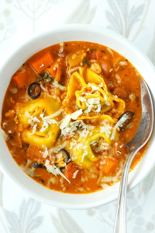 Tomato basil tortellini soup with pinto beans, chicken, diced carrot, and sliced black olives topped with grated cheese in a white round bowl with a silver spoon.