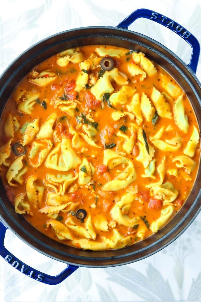 Creamy tomato basil tortellini soup in a blue Dutch oven on top of a floral grey napkin.