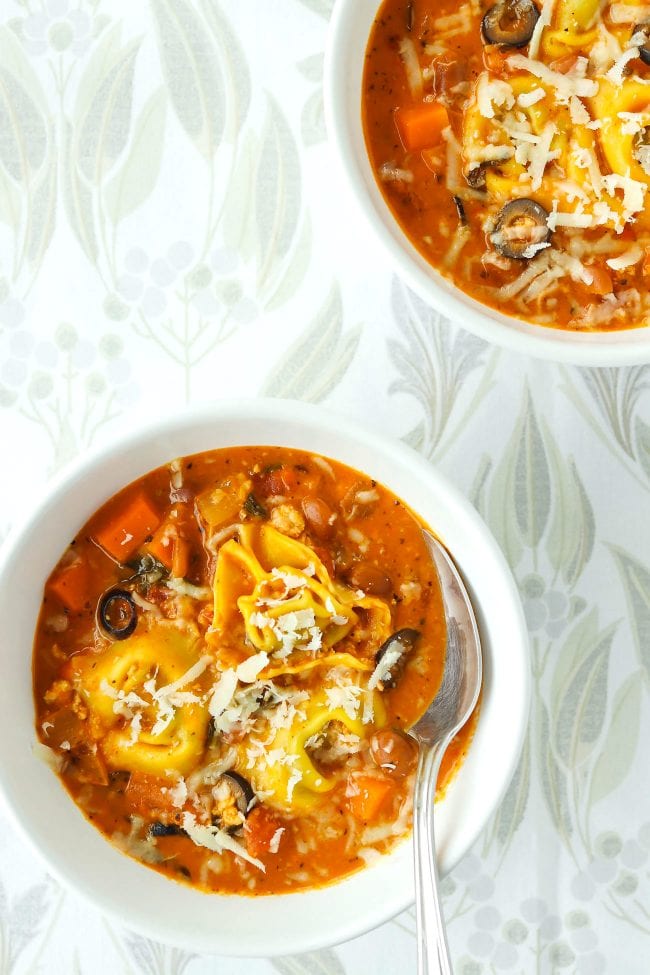 Two diagonally placed bowls of creamy tomato basil soup with tortellini, ground chicken, pinto beans, diced carrot, and sliced black olives topped with grated cheese. Bowl in front has a silver spoon in it.