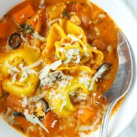 Close up of tomato basil tortellini soup with pinto beans, ground chicken, and sliced black olives topped with grated cheese in a white round bowl with a silver spoon.