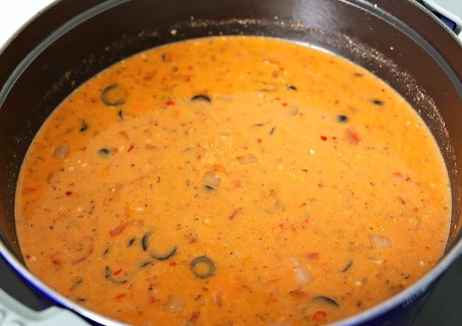 Tomato basil soup with sliced black olives, vegetables, and red chilies simmering in a Dutch oven on stovetop.