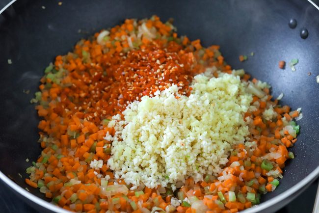 Minced garlic, ginger and red chilies on top of stir-fried chopped carrots, celery, onion, and spring onion whites in a smoking wok on the stovetop.