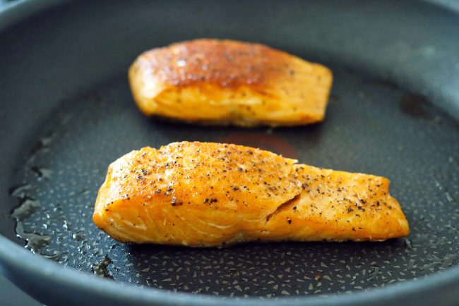 Two black pepper and salt seasoned salmon fillets pan-frying in a skillet with oil.