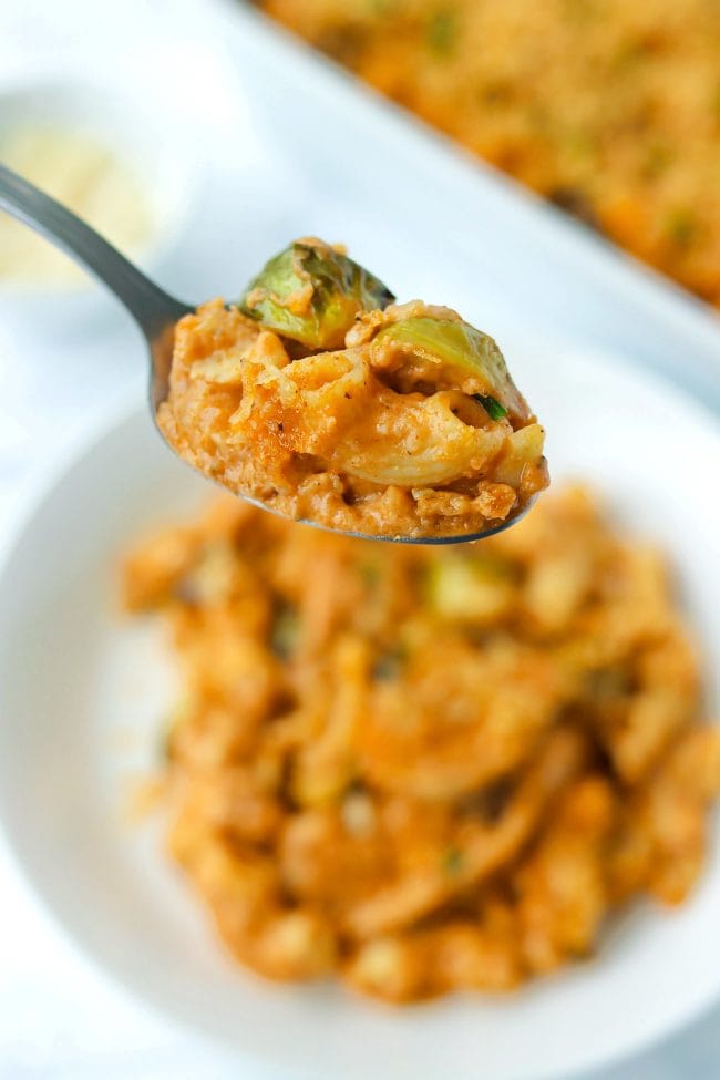 Spoon holding up a bite of harissa mac and cheese with brussels sprouts and a piece of chicken above a plate with mac and cheese. A large baking dish with mac and cheese and a small bowl with cheese shavings in the back.