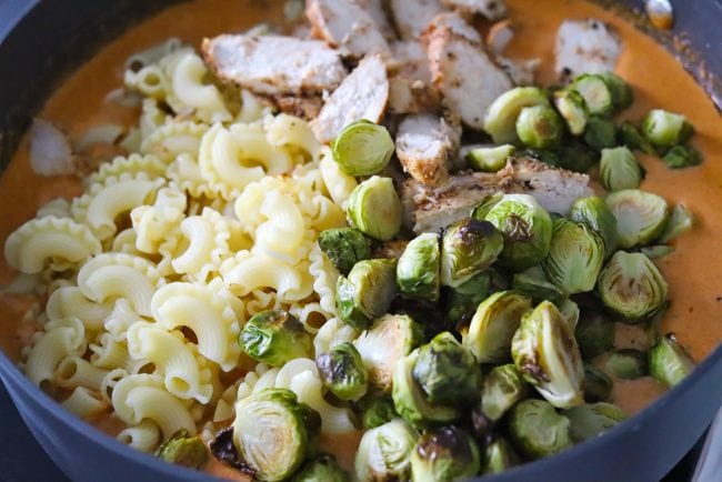 Unmixed pasta, roasted brussels sprouts, and chicken slices in a large black deep sauté pan with creamy spicy orange cheese sauce.