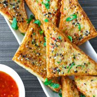Prawn Toast triangles on a long white plate garnished with chopped coriander and served with a small bowl of Thai Sweet Chili Sauce on the side.