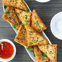 Spicy Prawn Toast triangles with Everything Bagel Seasonig on a long plate garnished with chopped coriander. Small bowl with Thai Sweet Chili Sauce and small plates on the side.