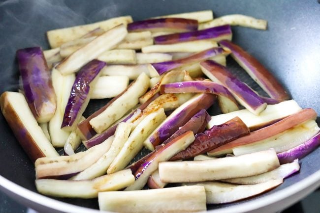 Eggplant strips softening and browning slightly in a wok on the stovetop.