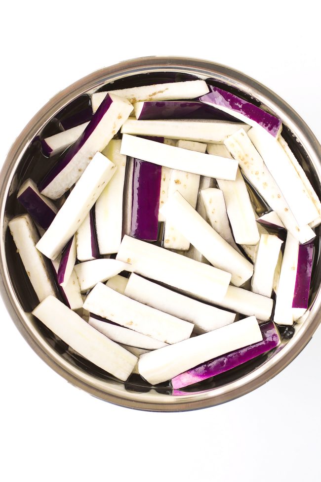 Eggplant cut into three inch strips soaking in a stainless steel bowl with water.