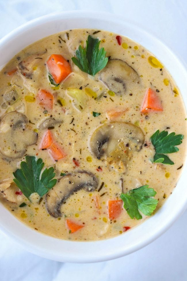 White round bowl with creamy soup with mushroom, chicken, carrots, celery, and garnished with parsley leaves and a drizzle of extra virgin olive oil.