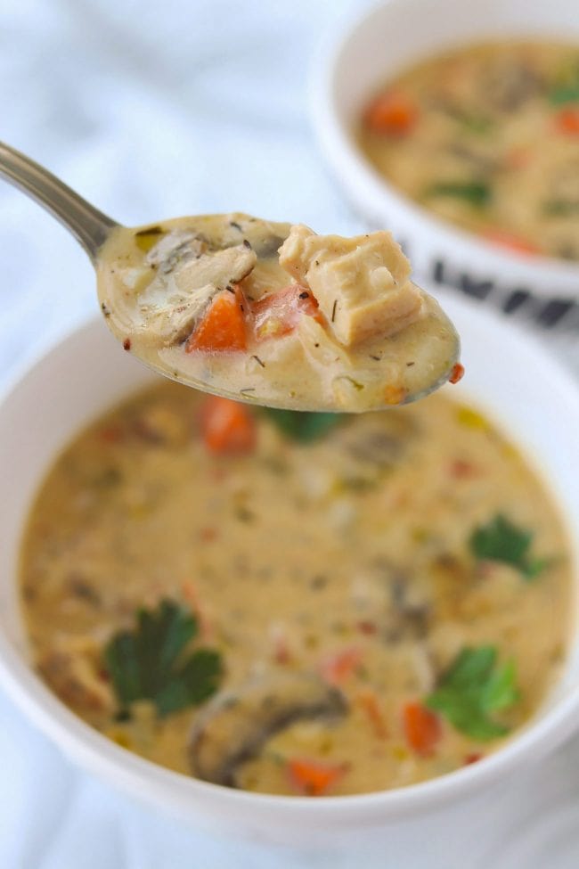 Spoonful of cream soup with mushroom, chicken, carrot and rice being held above a bowl with creamy mushroom and veggie soup and garnished with parsley leaves.