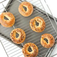 Two cheese bagels, three everything bagels, and a cheese and crushed red pepper bagel on a cooling rack on top of a baking tray.
