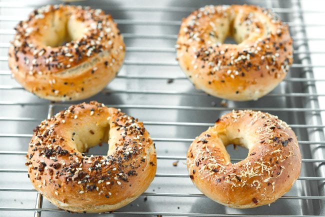Two grated cheese bagels, three everything bagels, and a crushed red pepper and cheese bagel on a cooling rack on top of a baking tray.