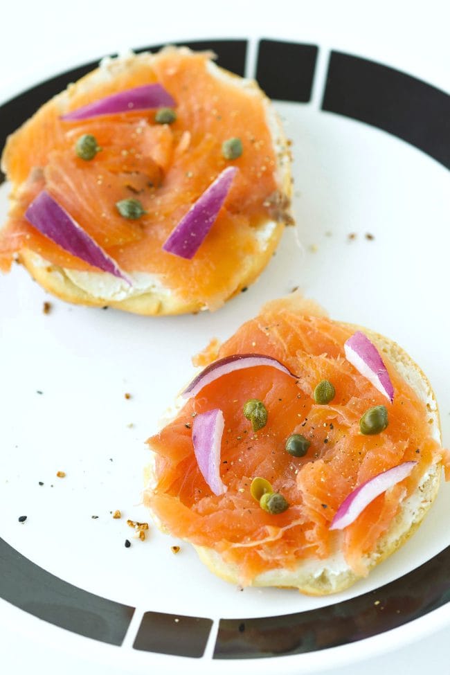 Bagel halves topped with cream cheese, smoked salmon, red onion slices, capers, and black pepper on a white and black plate.