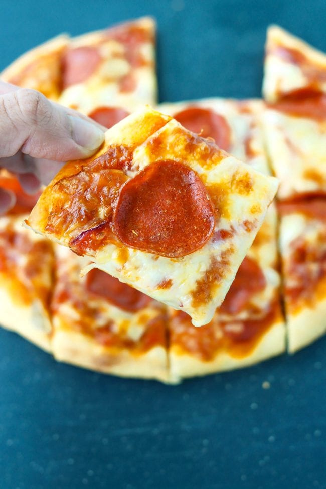 Hand holding up a pepperoni pizza square above a pepperoni pizza made with greek yogurt pizza dough.