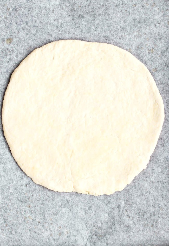 A round shaped unbaked pizza crust on parchment paper.