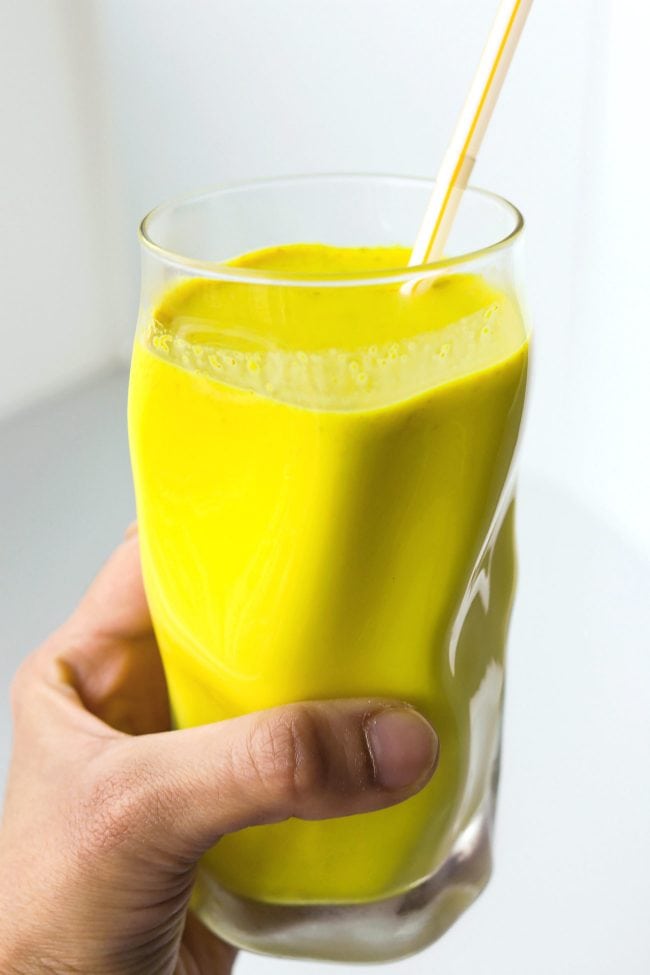 Hand holding up a glass with yellow turmeric milk with spices and a straw.