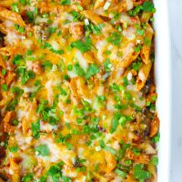 A large baking dish with Spicy BBQ Chicken Penne Pasta with corn, broccoli, melted cheese, and garnished with chopped coriander and spring onion.