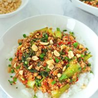 Ground pork and bok choy peanut sauce stir-fry on plate with rice, topped with sesame seeds, chopped peanuts, coriander, and spring onion. Serving bowl with the stir-fry and bowl with peanuts in the back.
