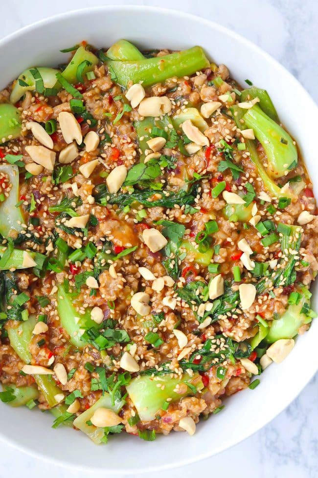 Ground pork and bok choy peanut sauce stir-fry in white round serving bowl topped with sesame seeds, chopped peanuts, coriander, and spring onion.