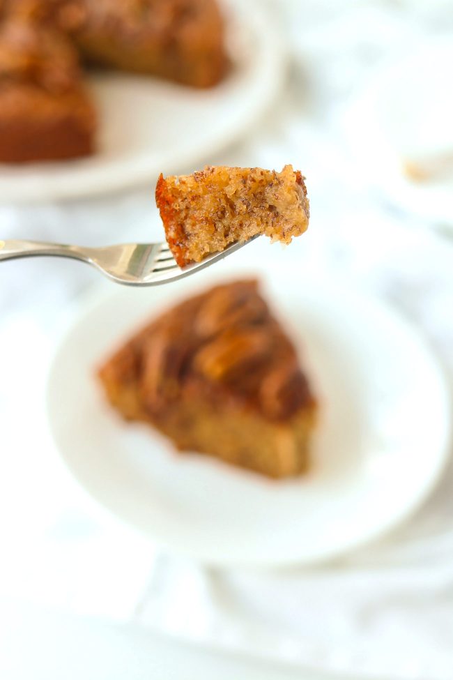 Fork holding up a bite of almond banana cake above a plate with a slice. Rest of the cake on a plate in the back.