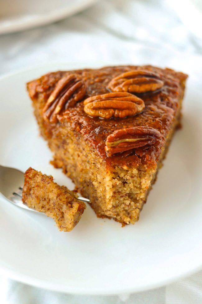 Close up of slice of almond meal banana cake topped with honey pecan glaze on a plate with a small bite of the cake on a fork.