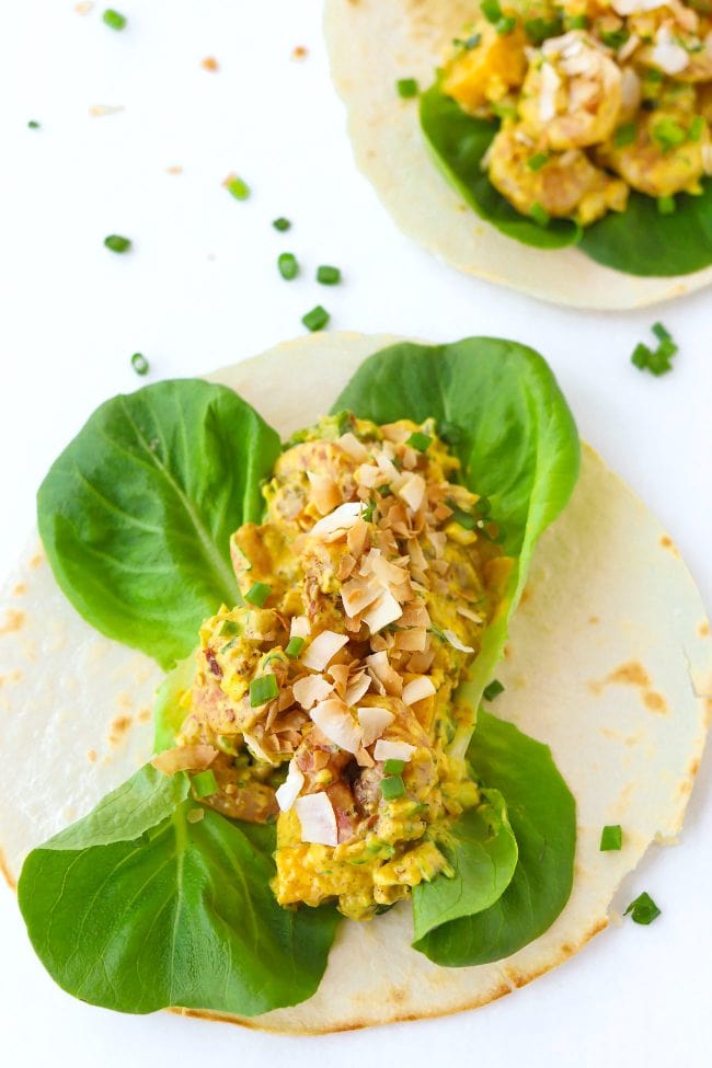 Two open-faced Cajun Shrimp Mango Tacos with butter lettuce and topped with toasted coconut flakes on a white background. Spring onion and coconut flakes scattered around the tacos.