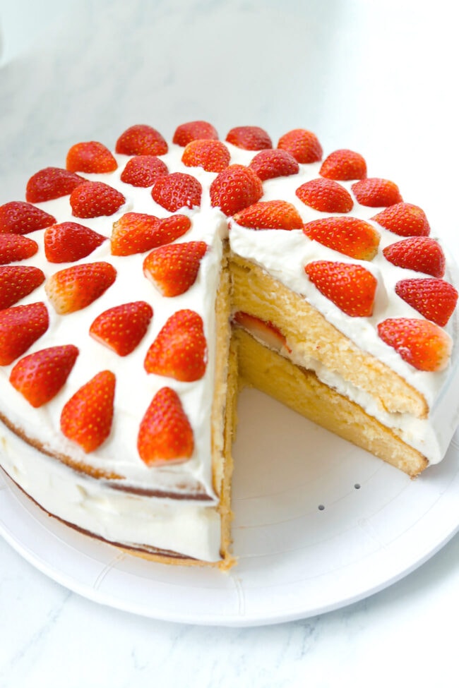 Top front angle view of strawberry cream layer cake on a platter with a slice cut out to show inside of cake.