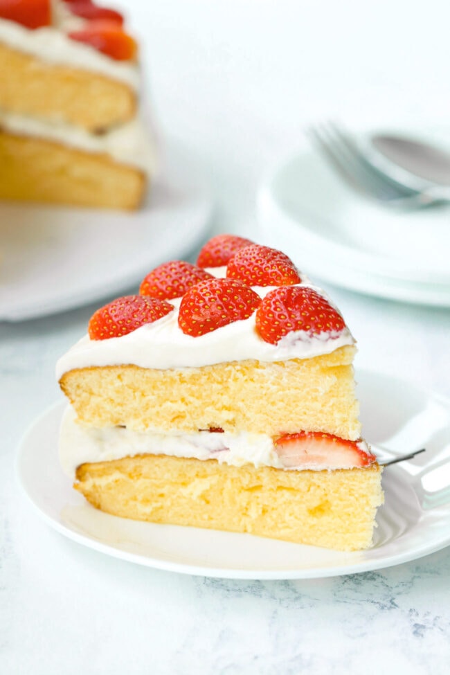Left side view of slice of strawberry cream layer cake on plate with fork. Rest of the cake on a platter and stacked plates with a fork behind.