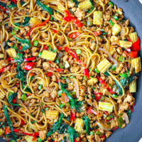 Overhead view of large wok with spaghetti tossed with baby corn, green beans, holy basil, ground chicken, chilies, onion, and garlic in a brown sauce. Text overlay "Thai Basil Chicken Spaghetti".