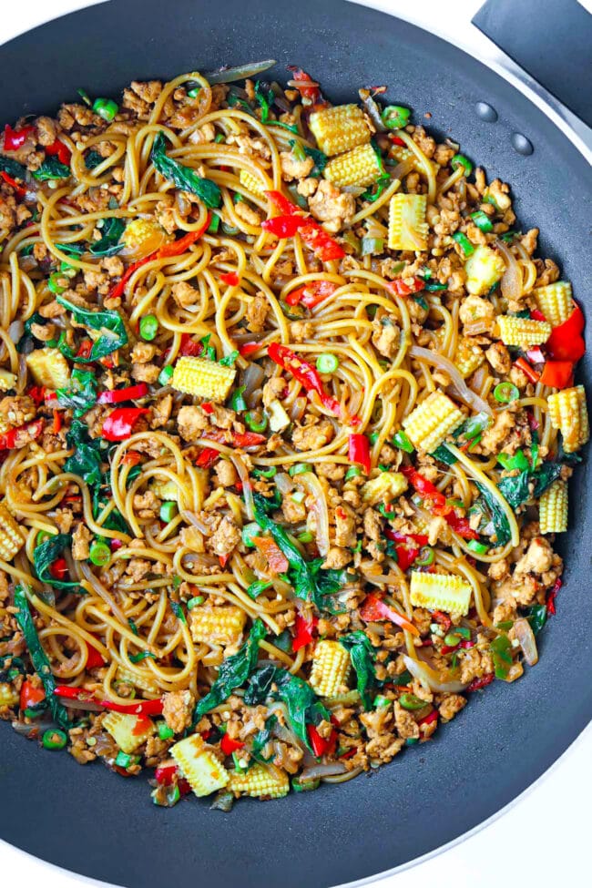 Top view of large wok with spaghetti tossed with ground chicken, red and green chilies, garlic, onion, baby corn, green beans, and holy basil in a brown sauce.