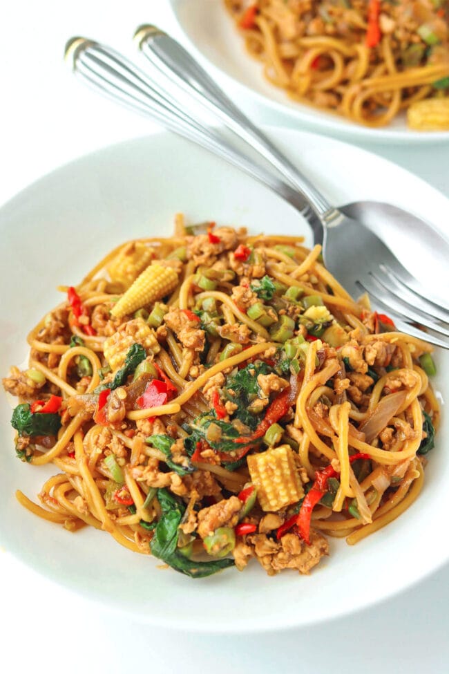 Two diagonally placed white round plates with spaghetti tossed with ground chicken, garlic, red and green chilies, onion, baby corn, green beans, and holy basil in a brown sauce. Fork and spoon on the top corner of the front plate.