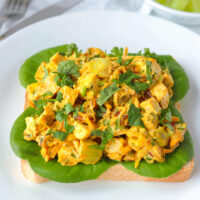 Front view of coronation chicken salad piled on a slice of bread with lettuce on a plate. Knife and fork and green grapes in a bowl in the back. Text overlay "Coronation Chicken Salad - curry chicken salad with dried apricots, grapes & pecans".