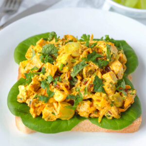 Front view of coronation chicken salad piled on a slice of bread with lettuce on a plate. Knife and fork and green grapes in a bowl in the back.
