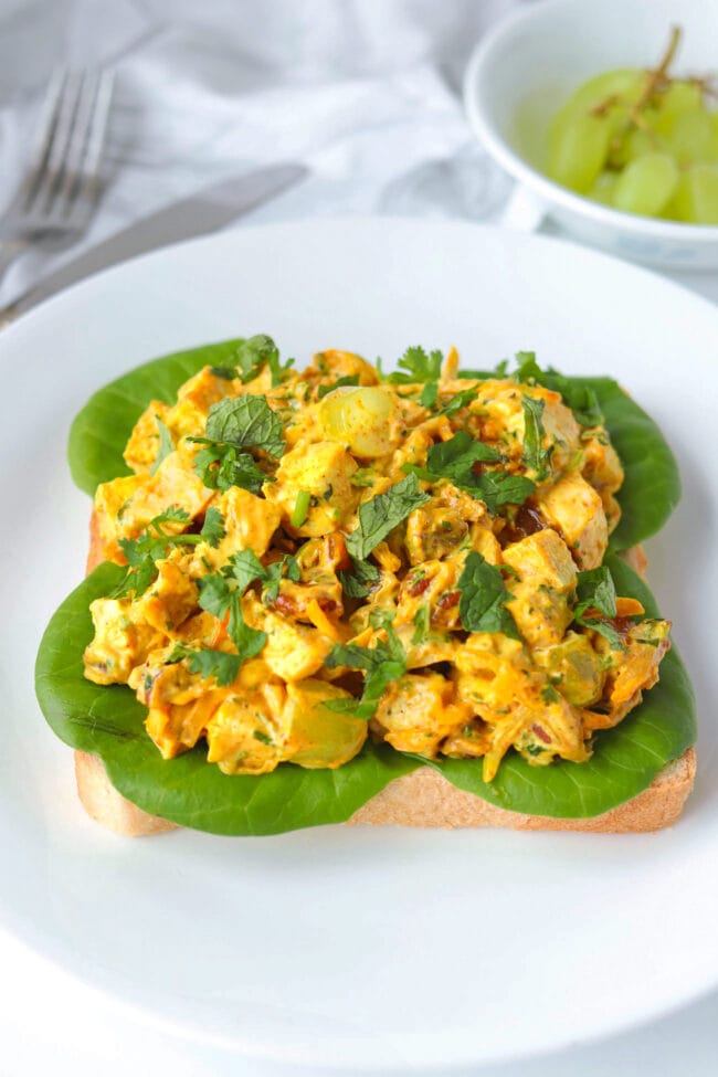 Front view of coronation chicken salad on a slice of bread with lettuce on a plate. Utensils and green grapes in a bowl in the back.