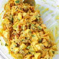 Tossed coronation chicken salad ingredients in a large mixing bowl with a spoon. Text overlay "Coronation Chicken Salad".