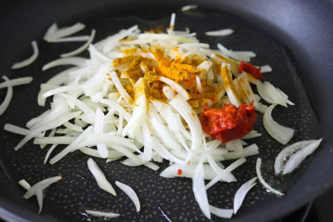 Sliced onion topped with curry powder, turmeric, and tomato paste in a pan with oil.
