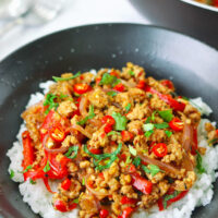 Front view of ground pork stir-fry garnished with mint leaves and coriander on rice in a bowl. Wok with the stir-fry in the back. Text overlay "Lemongrass Pork Stir-fry".