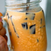 Hand holding up a mason mug with Thai milk tea, cubed grass jelly, and ice with a straw and tall spoon. Text overlay "Thai Iced Milk Tea with Grass Jelly".